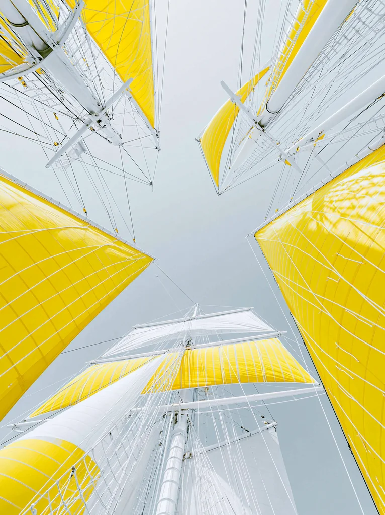 White ship masts with lemon yellow sails seen straight up around a pale gray sky